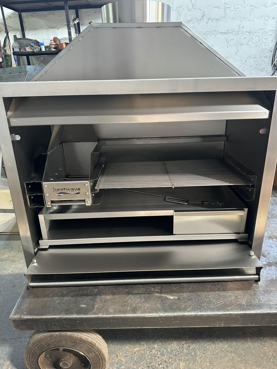 Interior of an open stainless steel braai showing the ember maker, grill grid and slide-out ash drawer